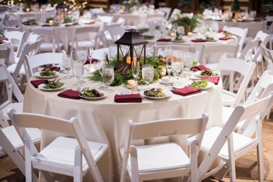 Several large circular tables with white tablecloths surrounded by white folding chairs. The tables are set with plates of salad at each seat, a nd a red napkin at each seat. In the middle of teh table is a large decorative setup with a candle and some greenery