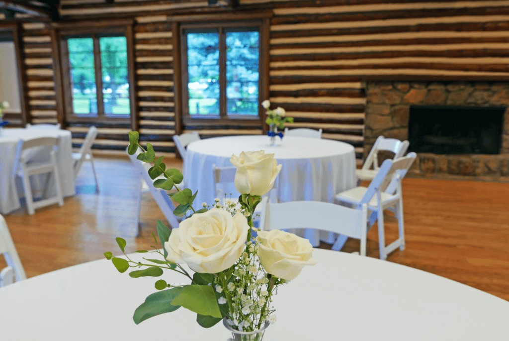 Several white circular tables inside the Inn at Hudson Gardens. The tables have vases with white roses on them and the tables are surrounded by white folding chairs