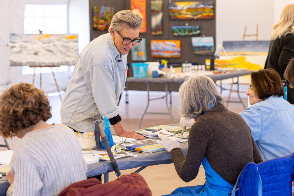 Artist instructing student on painting with display of artworks in the background