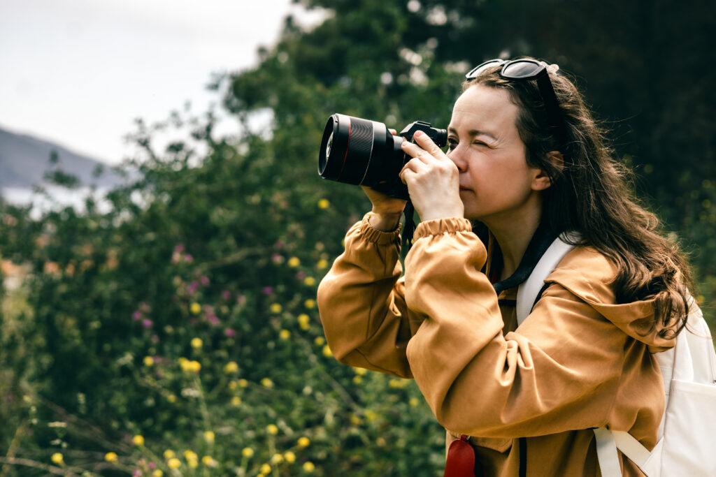 Young woman with long hair and backpack taking photo with professional camera outdoors, enjoying nature, travel, and photography in scenic green landscape with soft natural light.