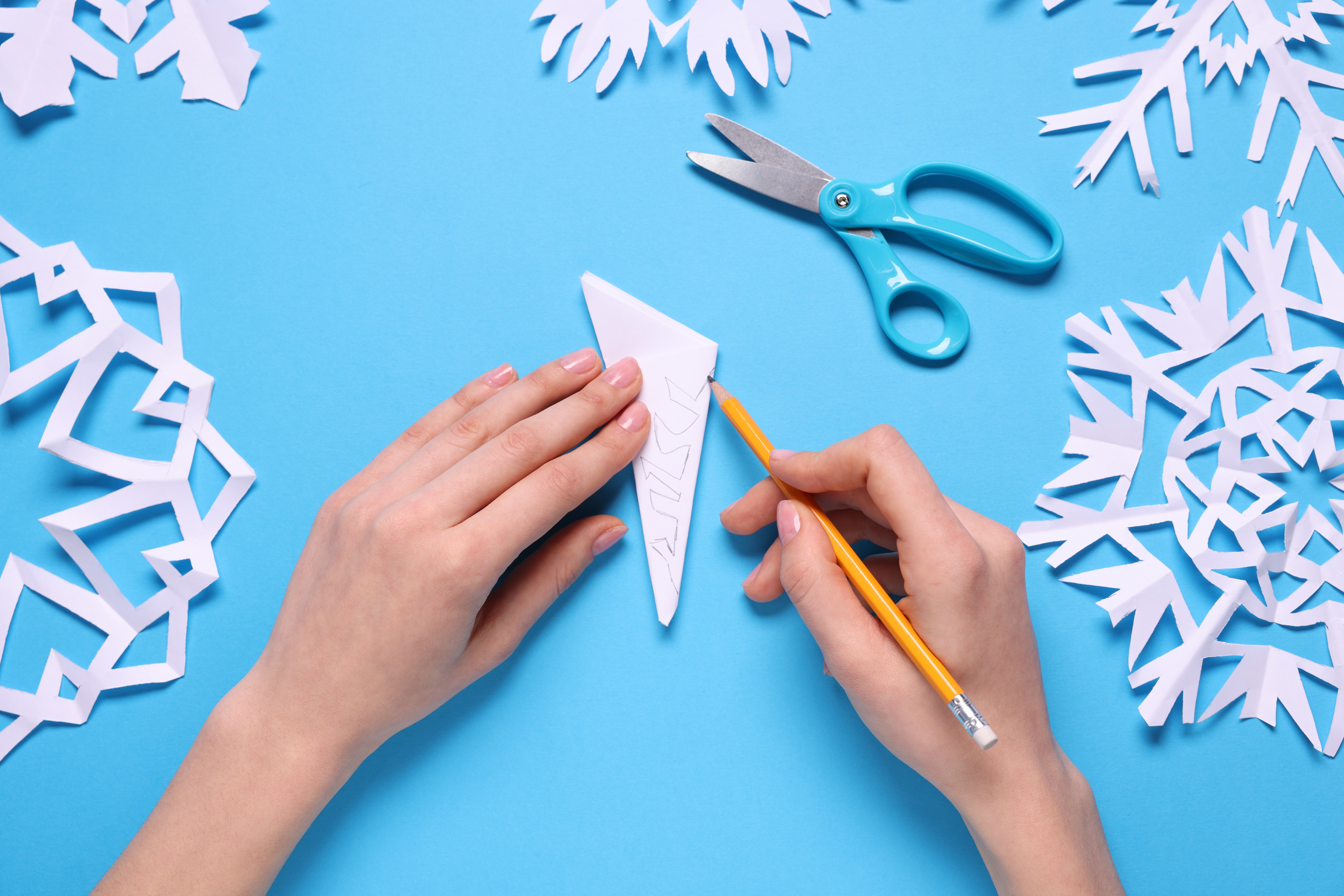 Woman making paper snowflake on light blue background, top view